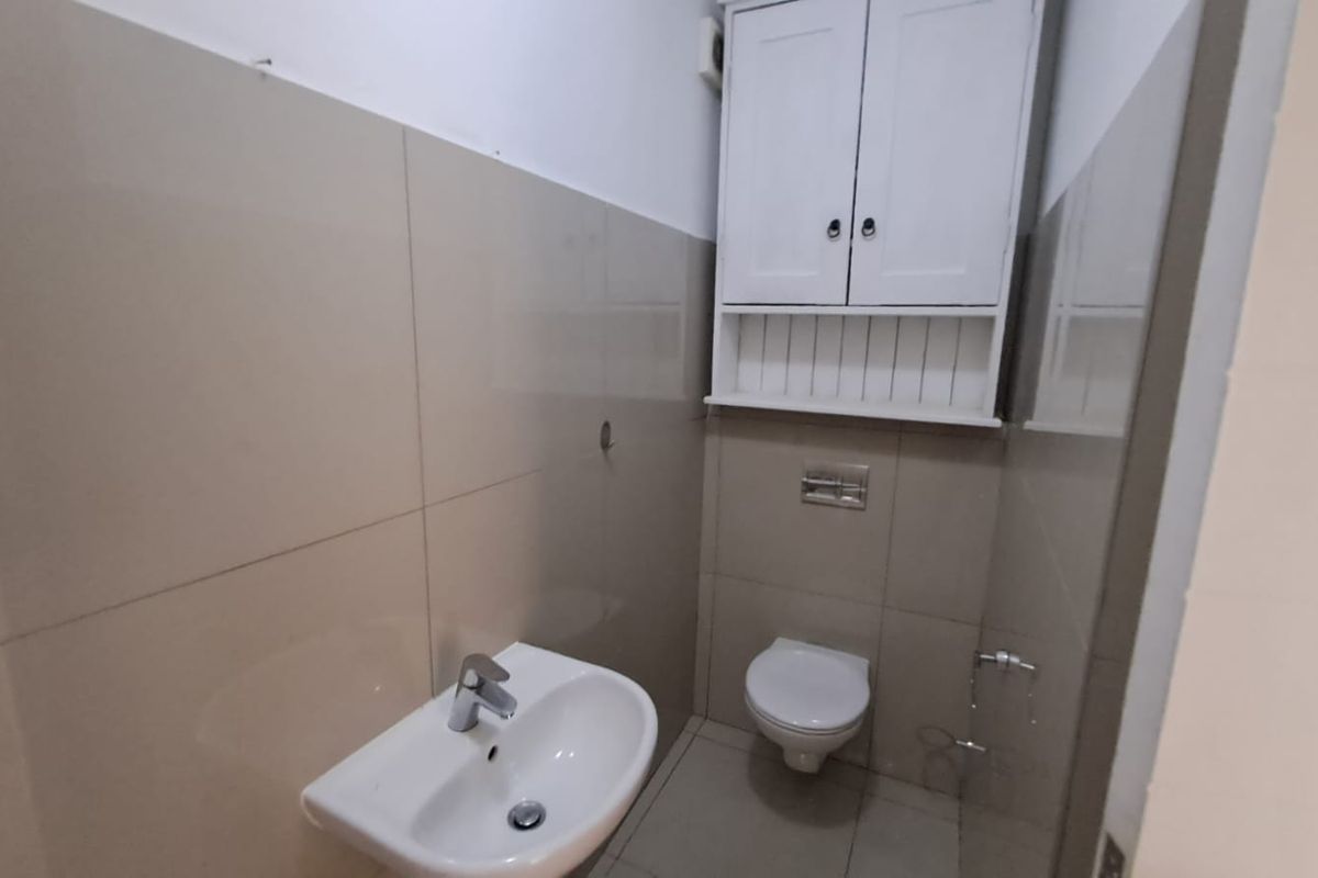 A compact bathroom featuring neutral wall tiling, a wall-mounted basin, a wall-hung toilet, and a white overhead storage cabinet.