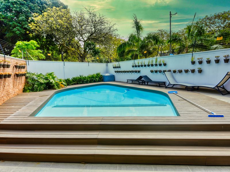 A well-maintained outdoor pool area featuring modern timber decking, secure boundary walls with electric fencing, and established garden surroundings.