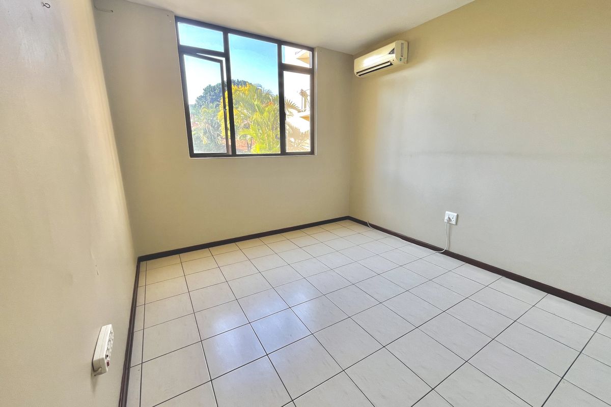 A bright, empty bedroom featuring tiled flooring, a large window, and a wall-mounted air conditioning unit.