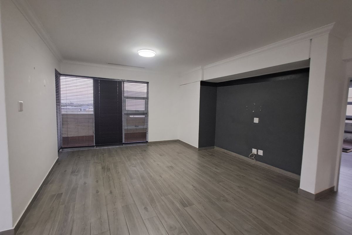 A spacious, empty living room featuring modern grey wood-look flooring, a dark accent wall, and large windows fitted with horizontal blinds.