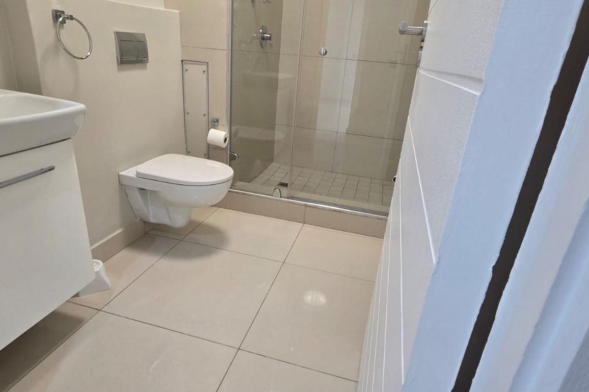 A modern, clean bathroom featuring a wall-hung toilet, vanity basin, and a glass-enclosed shower with neutral floor tiling.