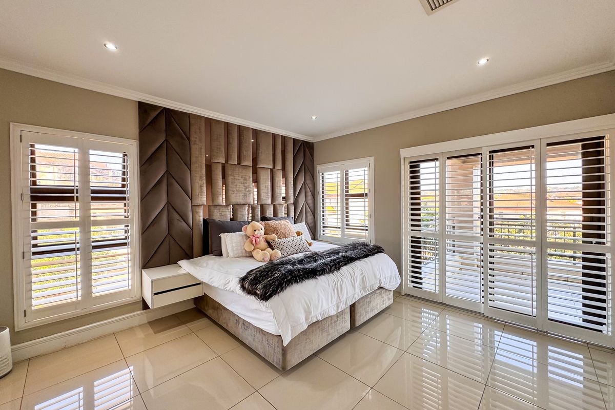 Spacious bedroom featuring polished porcelain floor tiles, custom upholstered headboard wall, recessed downlighting, and plantation shutters opening onto a private balcony.