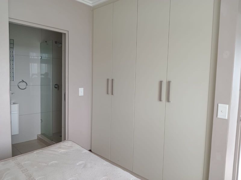 A clean bedroom featuring neutral built-in cupboards and an adjoining en-suite bathroom with a modern glass shower enclosure and vanity.
