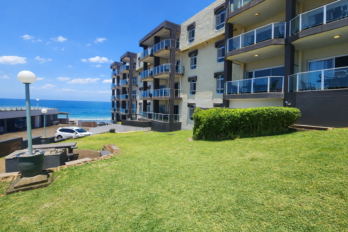 Exterior view of a multi-story apartment complex featuring private balconies with glass railings, overlooking a landscaped lawn area with ocean views.