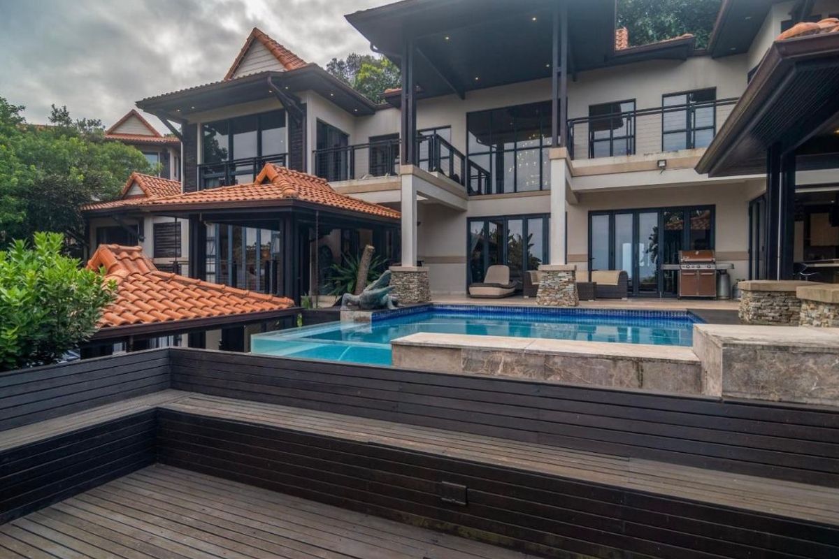 A luxurious multi-level rear exterior featuring a modern swimming pool, expansive timber decking, and covered patio areas with architectural detailing.