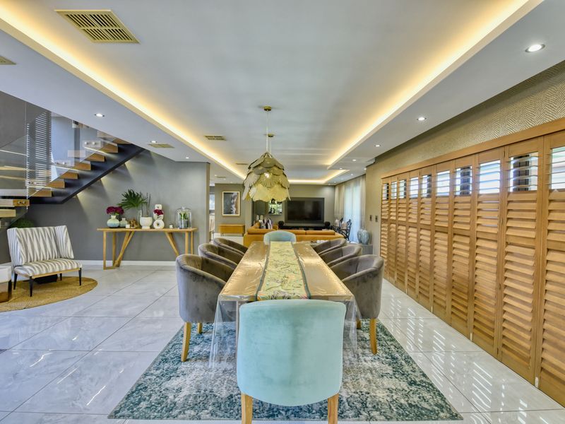 Spacious open-plan dining and living area featuring high-end finishes, recessed cove lighting, a modern floating staircase with glass balustrade, and elegant timber plantation shutters.