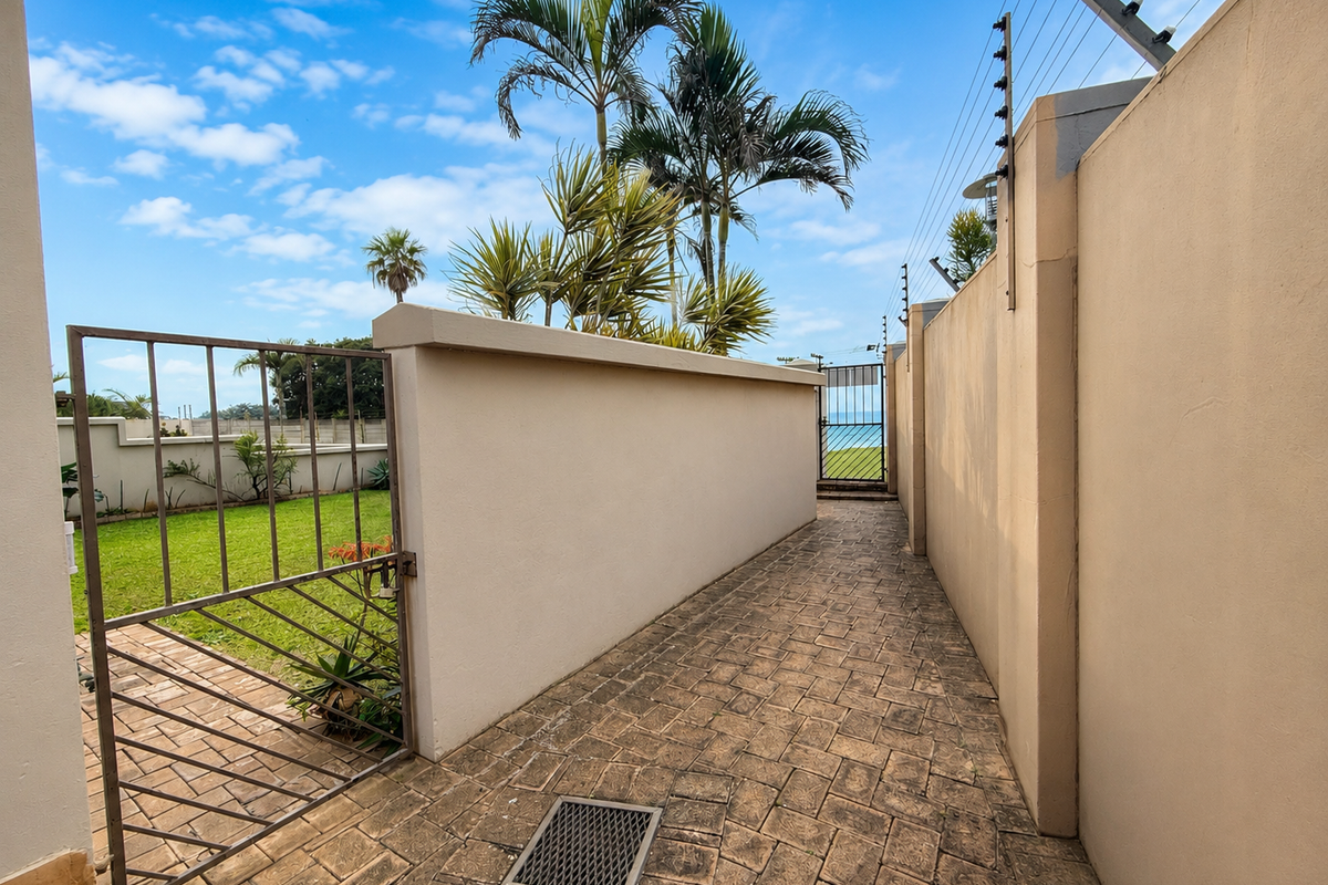A secure rear exterior walkway featuring paved flooring, a metal gate, boundary walls with electric fencing, and a small lawn area.