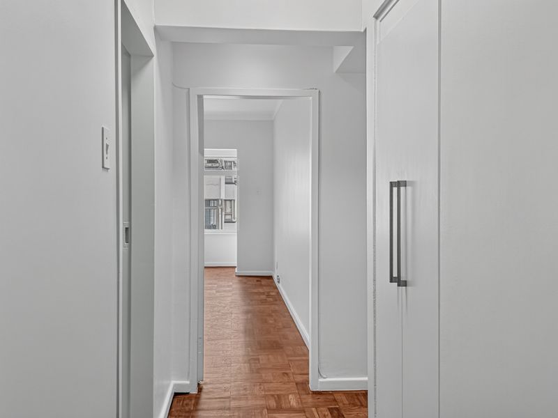 A bright, clean hallway featuring classic parquet flooring and built-in storage cabinetry, leading through to an adjacent room.