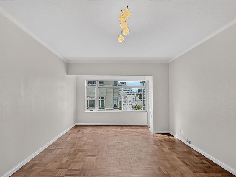 A bright, empty living room featuring classic parquet flooring, neutral walls, and a large window offering an urban view.