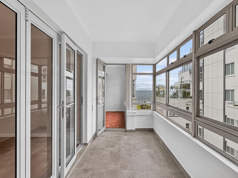 A bright, enclosed balcony featuring modern tiled flooring and expansive aluminium-framed windows offering pleasant neighbourhood and partial sea views.