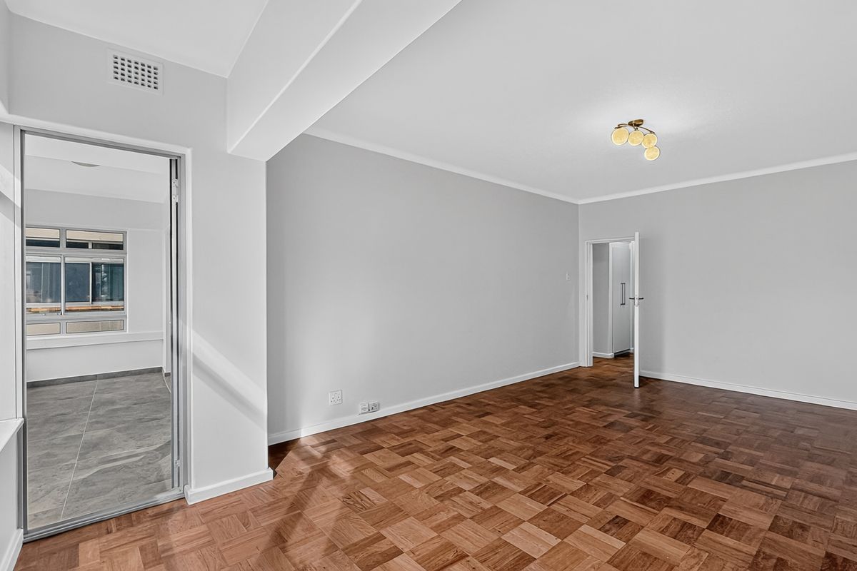 A bright and spacious living area featuring well-maintained parquet flooring, neutral walls, and direct access to an adjoining room.