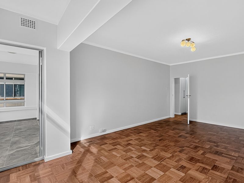 A bright and spacious living area featuring well-maintained parquet flooring, neutral walls, and direct access to an adjoining room.