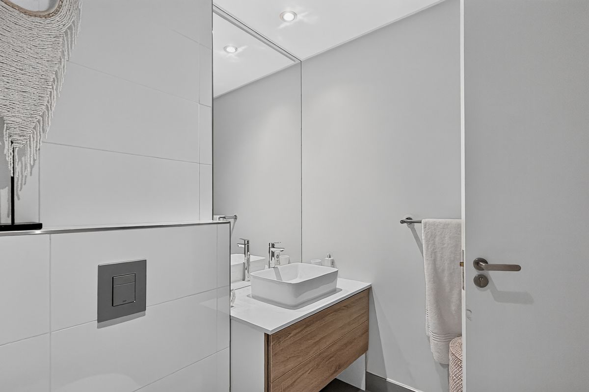 Modern bathroom featuring a sleek vanity with a vessel sink, wall-to-wall mirror, and wall-mounted toilet flush plate in a clean, contemporary finish.
