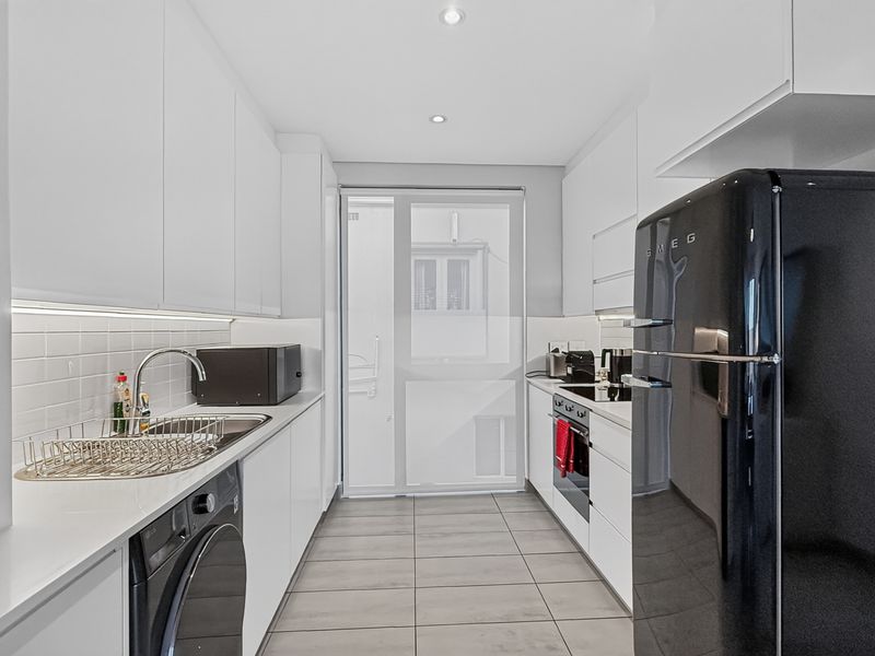 Modern galley-style kitchen featuring sleek white cabinetry, subway tile backsplash, integrated appliances, and tiled flooring with direct access to an outdoor area.