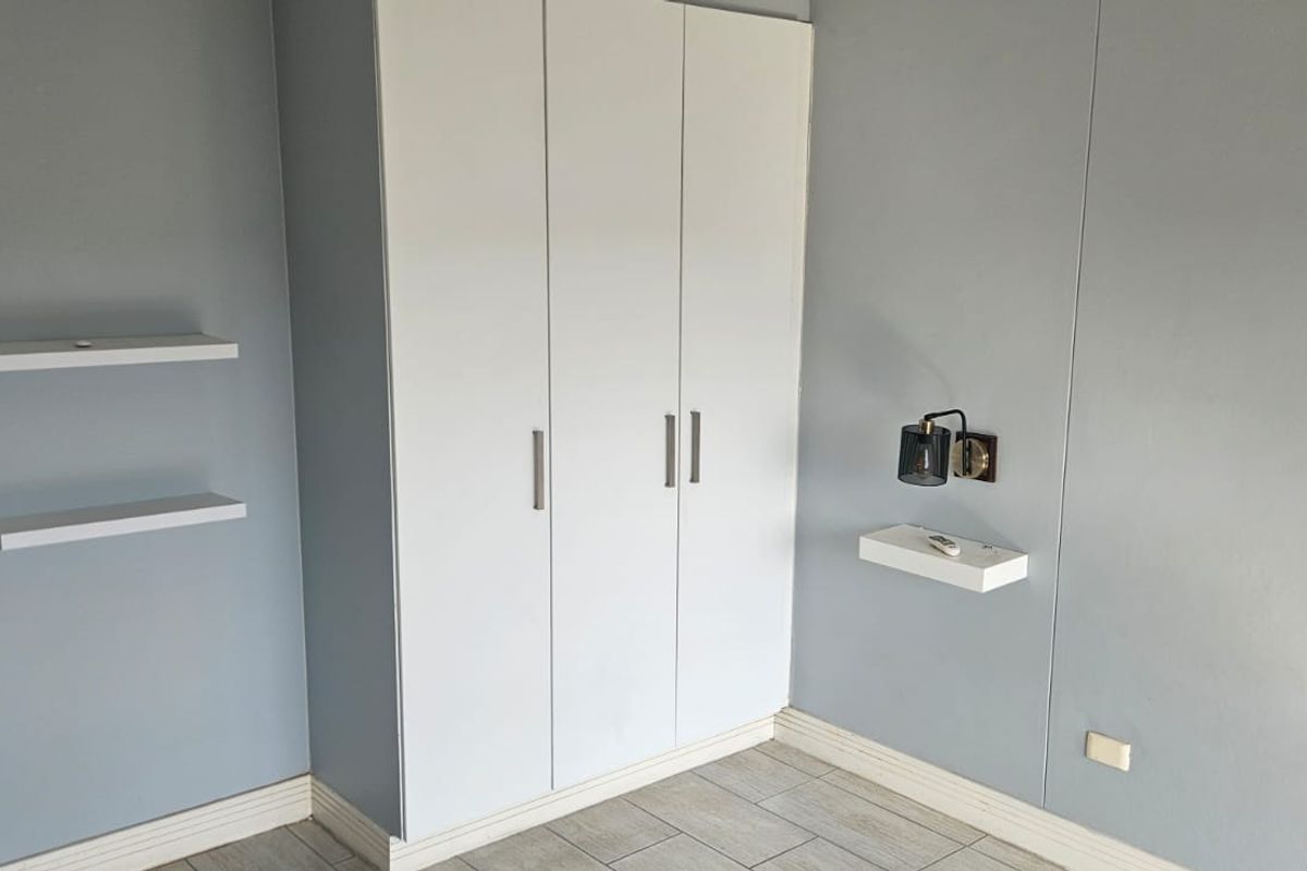 A clean, modern bedroom featuring built-in white wardrobes, floating shelving, and neutral grey walls with tiled flooring.