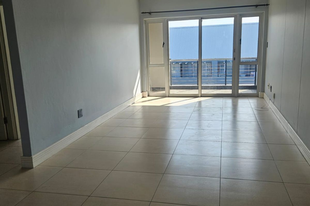 An empty, light-filled living room featuring neutral tiled flooring and a large sliding glass door providing access to an outdoor balcony.