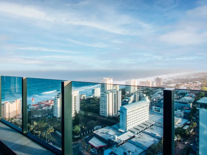 A high-rise balcony featuring a modern glass balustrade with an expansive panoramic view of the coastline and urban cityscape.