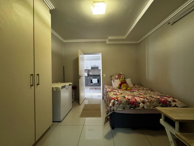 A bedroom featuring tiled flooring, built-in cupboards, and decorative ceiling cornicing, with a view through to the adjacent kitchen area.