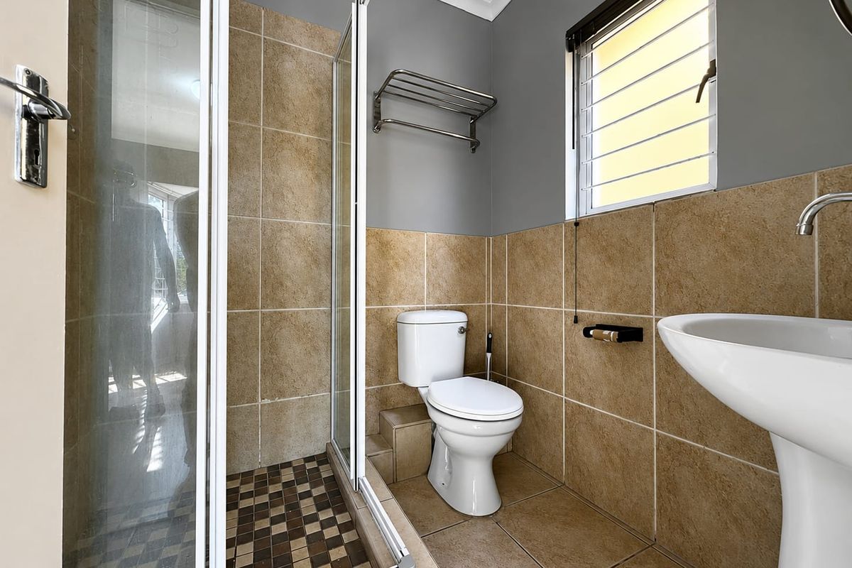 A clean, functional bathroom featuring a shower enclosure, pedestal basin, toilet, and neutral tiled finishes with a window for natural light.
