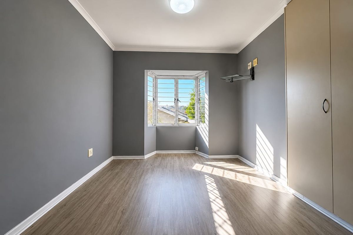 A bright, empty bedroom featuring modern grey walls, light wood-look laminate flooring, a built-in wardrobe, and a bay window with security bars.