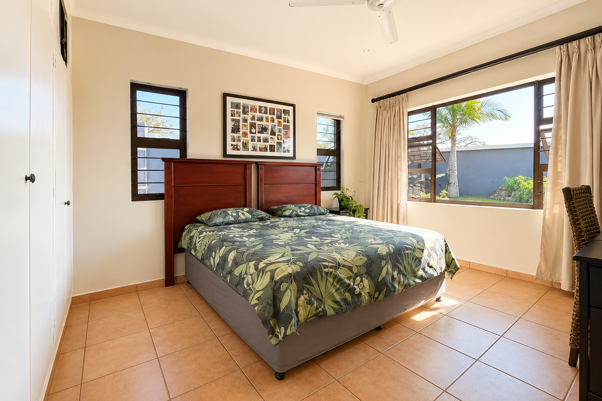 A bright bedroom featuring tiled flooring, built-in cupboards, a ceiling fan, and large windows providing natural light and garden views.