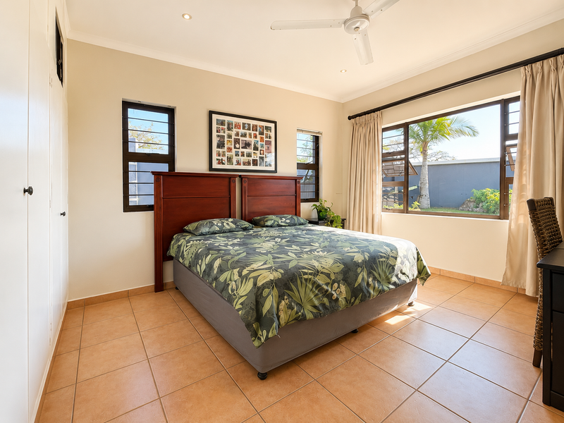 A bright bedroom featuring tiled flooring, built-in cupboards, a ceiling fan, and large windows providing natural light and garden views.