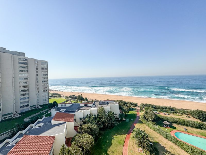Elevated coastal view overlooking the beach, surrounding apartment complex, landscaped gardens, and communal swimming pool area.