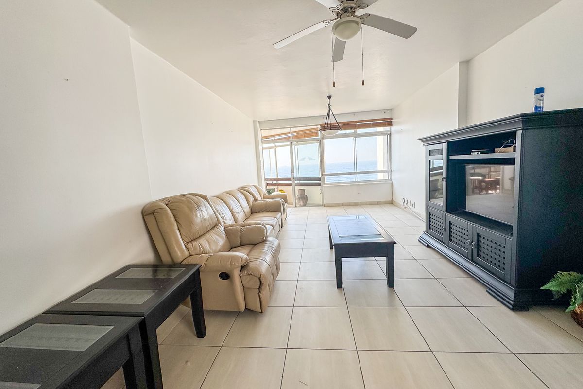 Spacious living room featuring neutral tiled flooring, a ceiling fan, and large windows providing natural light and ocean views.