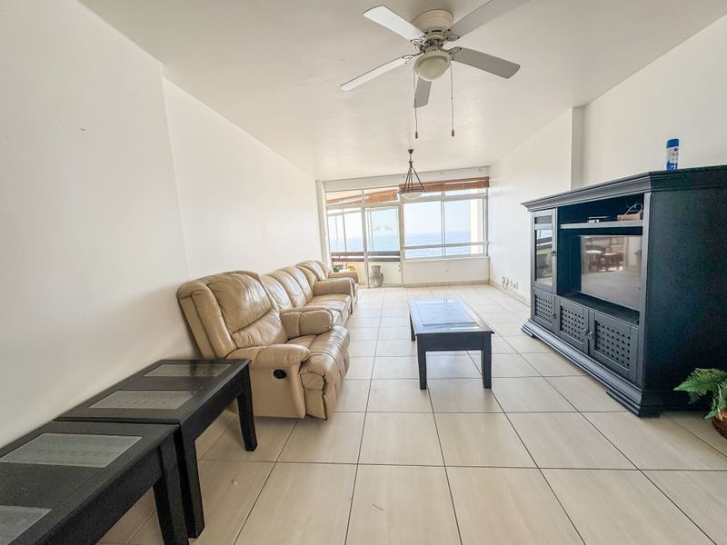 Spacious living room featuring neutral tiled flooring, a ceiling fan, and large windows providing natural light and ocean views.