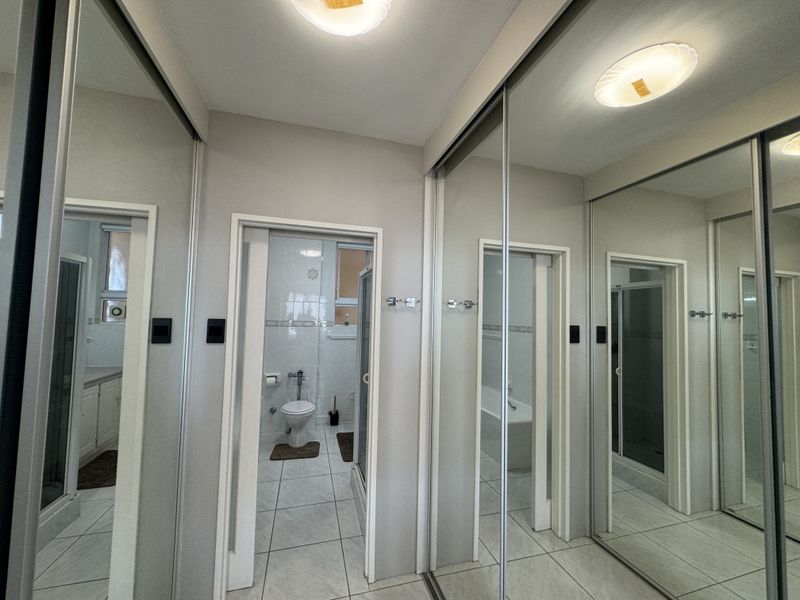A clean hallway featuring floor-to-ceiling mirrored sliding wardrobe doors and neutral tiled flooring, providing access to the adjacent bathroom.