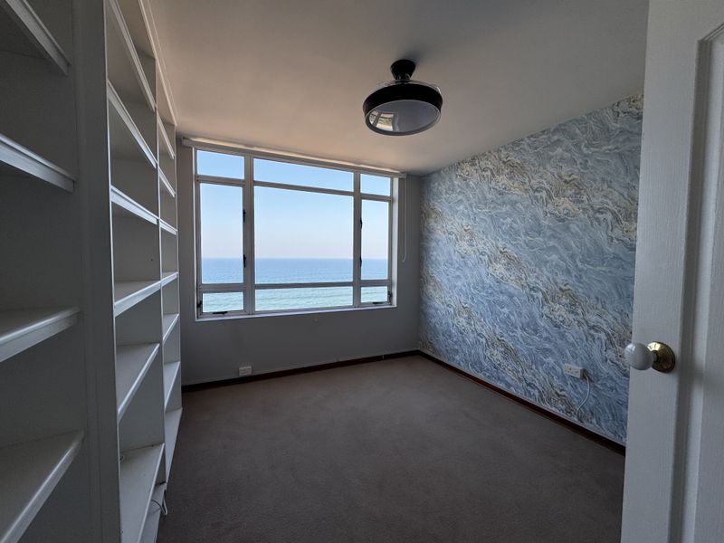 A bright, empty bedroom featuring built-in shelving, decorative wallpaper, and a large window offering expansive ocean views.