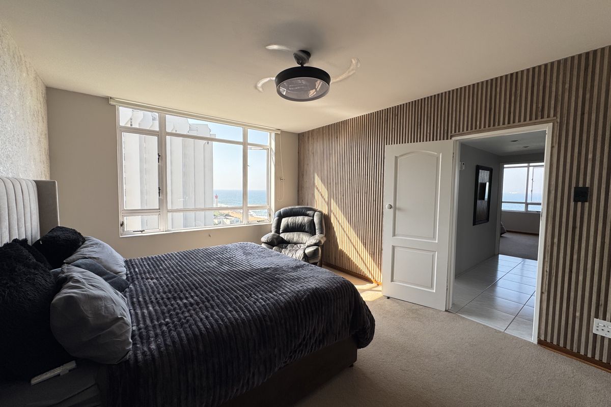 Spacious bedroom featuring large windows with ocean views, modern slatted wood wall paneling, and a ceiling fan, leading into an adjacent hallway.