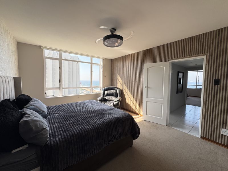 Spacious bedroom featuring large windows with ocean views, modern slatted wood wall paneling, and a ceiling fan, leading into an adjacent hallway.