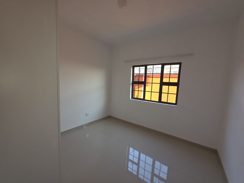 A clean, empty bedroom featuring neutral tiled flooring and a window with security bars, offering a bright and versatile living space.