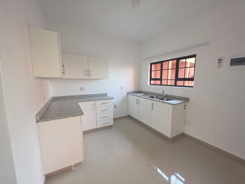 A clean, empty kitchen featuring white cabinetry, granite countertops, a double stainless steel sink, and tiled flooring.