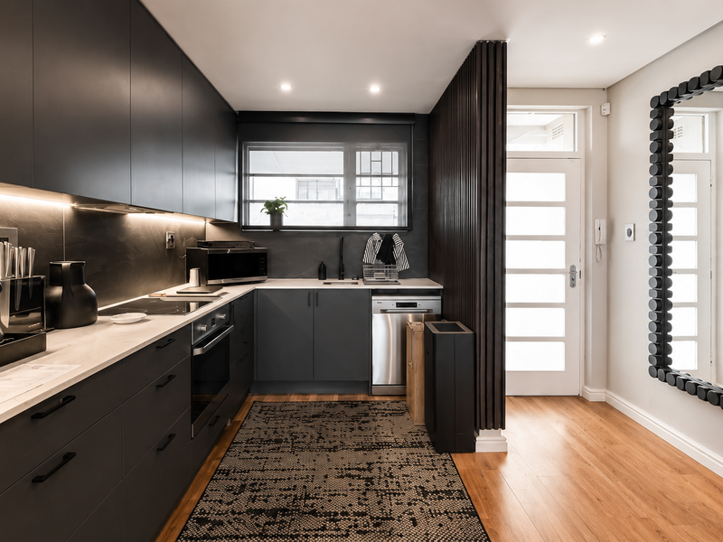 Modern galley-style kitchen featuring matte dark cabinetry, stone countertops, integrated appliances, and warm timber flooring, seamlessly connected to the entrance area.