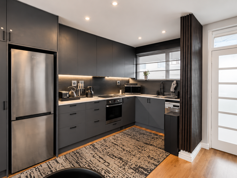 Modern kitchen featuring sleek dark cabinetry, integrated appliances, timber flooring, and a stylish slatted wood feature wall with direct exterior access.