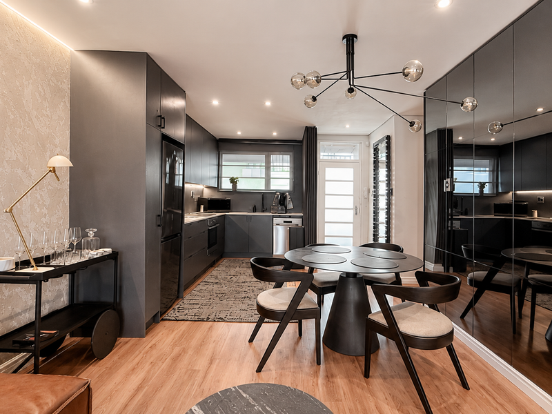 Modern open-plan kitchen and dining area featuring sleek dark cabinetry, integrated appliances, mirrored wall accents, and warm wood-look flooring.