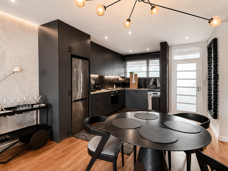 Modern open-plan kitchen and dining area featuring sleek matte black cabinetry, integrated appliances, timber flooring, and a stylish textured feature wall.