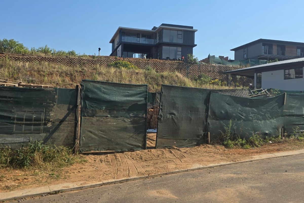 A vacant residential plot featuring a retaining wall, currently enclosed by temporary site fencing with surrounding neighborhood development visible.