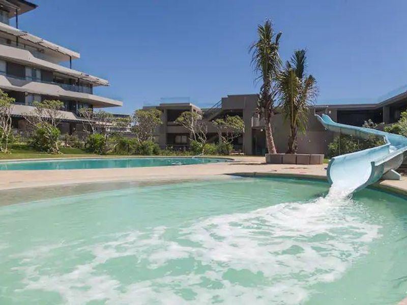 Communal swimming pool area featuring a water slide, paved deck, and landscaped surroundings within the apartment complex.