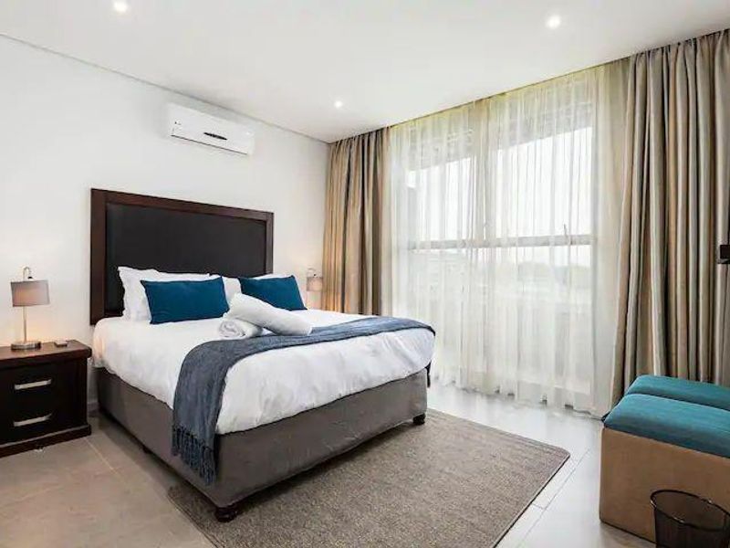 A well-maintained bedroom featuring tiled flooring, recessed lighting, and a wall-mounted split-system air conditioner with large windows covered by floor-to-ceiling curtains.