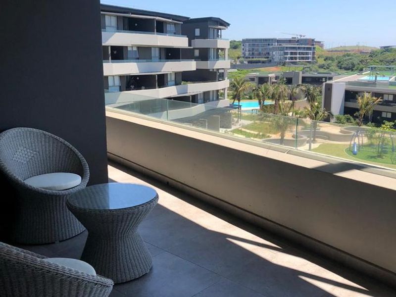 A spacious covered balcony featuring modern tiled flooring and a glass balustrade, offering pleasant views of the surrounding apartment complex and landscaped grounds.