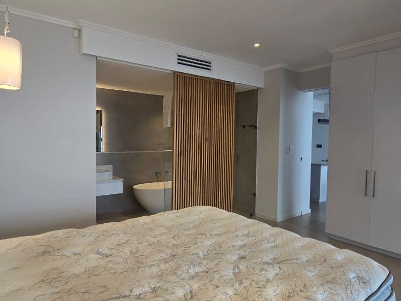 Modern bedroom featuring built-in wardrobes, ducted air conditioning, and an open-plan ensuite with a freestanding bathtub and stylish timber slat privacy screen.