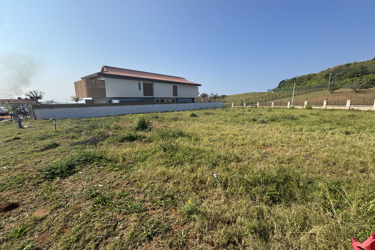 A view of a vacant residential plot featuring a sloped grassy landscape, enclosed by a perimeter wall and boundary fencing.