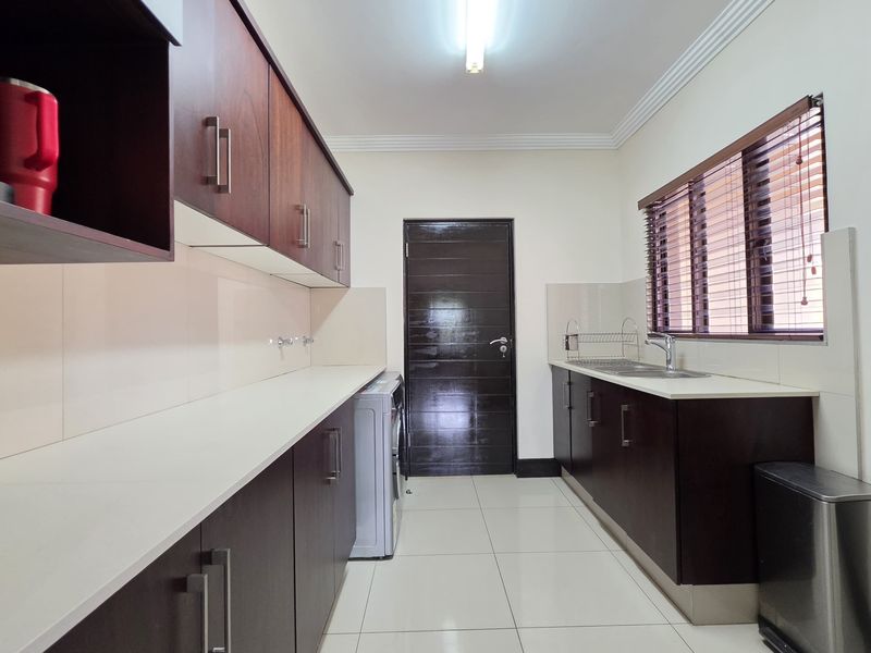 A functional scullery featuring dark wood cabinetry, light countertops, tiled flooring, and a stainless steel sink with convenient access to the main living area.