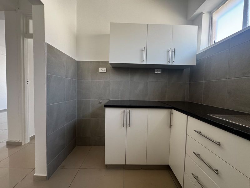 A clean, functional kitchen featuring white built-in cabinetry, dark granite countertops, grey tiled splashbacks, and tiled flooring in a well-lit, compact layout.