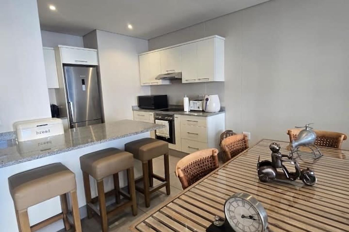 Modern open-plan kitchen and dining area featuring sleek white cabinetry, granite countertops, integrated appliances, and neutral tiled flooring throughout.