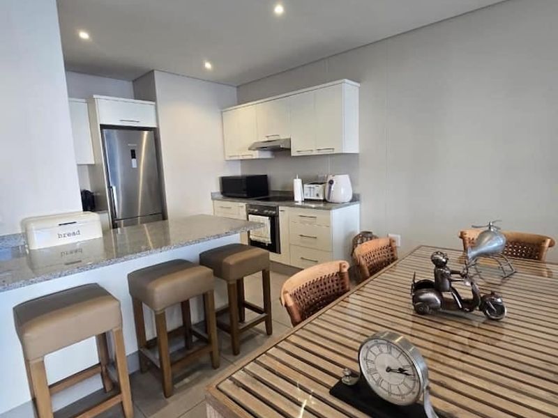 Modern open-plan kitchen and dining area featuring sleek white cabinetry, granite countertops, integrated appliances, and neutral tiled flooring throughout.