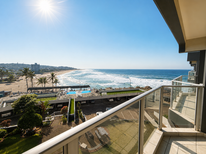 A spacious balcony featuring a modern glass balustrade and tiled flooring, offering expansive, elevated views of the coastline and ocean.